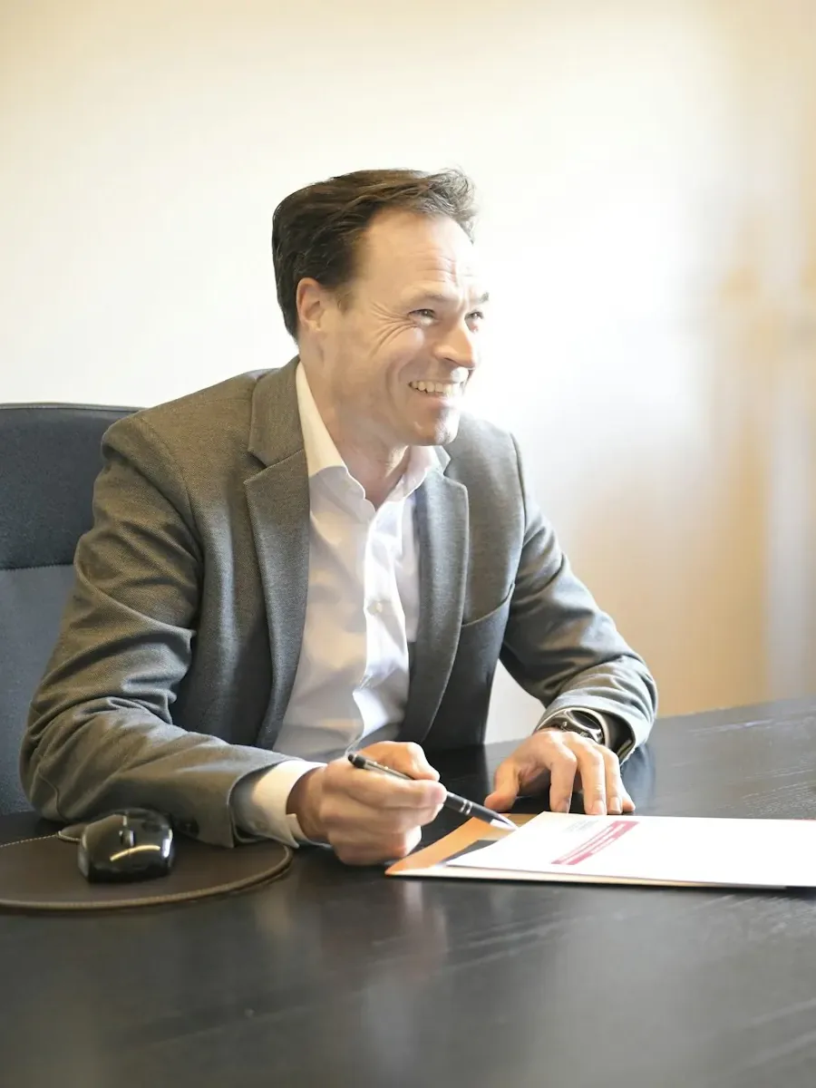 Un homme sourit à son bureau, stylo en main, devant un document.